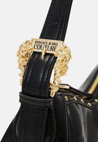 Czarna skórzana torebka z ozdobną złotą klamrą z napisem "Versace Jeans Couture". Widoczne suwaki i ozdobne ćwieki.