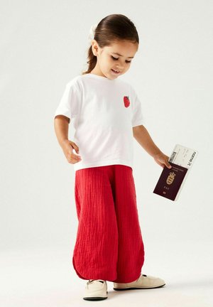 Ragazza giovane con camicia bianca con stampa di fragole e pantaloni rossi che tiene un passaporto e una carta d'imbarco, in piedi su uno sfondo bianco.