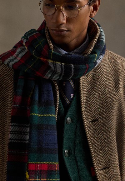 Cachecol em mistura de lã com padrão xadrez em verde, azul-marinho e vermelho, enrolado em volta de um casaco de espinha de peixe, sobreposto a um suéter verde e uma gravata estampada.