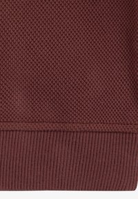 Tessuto lavorato a maglia bordeaux con un motivo testurizzato e orlo a coste. La trama è densamente lavorata, offrendo un aspetto sostanziale e caldo.