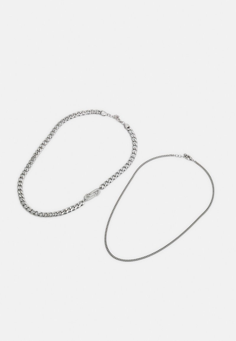 Duas correntes de prata: uma com um fecho robusto e a outra uma corrente fina e delicada. Ambas possuem um acabamento metálico suave e um brilho subtil.