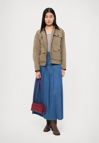 Jachetă khaki cu patru buzunare, top gri, fustă lungă din denim albastru, bocanci maronii și o geantă roșie de umăr. Texturi netede și culori neutre.