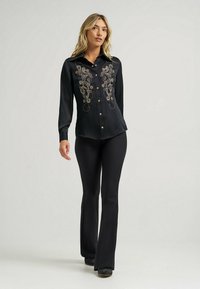Chemise en satin noir avec une broderie dorée complexe sur le devant, fermeture à boutons et manches longues, associée à un pantalon noir évasé et des bottes.