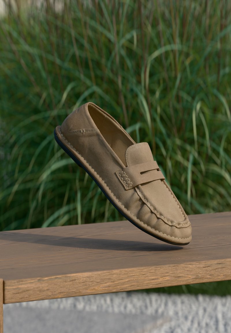 Mocassin en daim beige avec détails cousus, présenté en suspension au-dessus d'un banc en bois, avec un fond d'herbe verte.