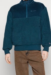 Teal fleecetröja med en snodd, texturerad övre del; har en dragkedja i kragen, elastiska ärmavslut och en logotyp på bröstet.