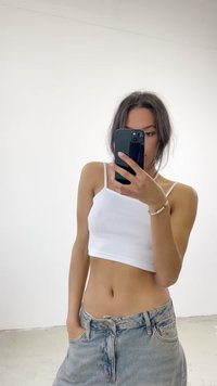 Top corto bianco a coste e gonna di denim azzurra con vestibilità rilassata. La modella posa di fronte a uno sfondo bianco semplice, tenendo uno smartphone.