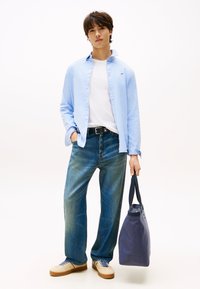 Šviesiai mėlyna marškinėliai su sagomis virš balto marškinėlio, derinami su laisvais mėlynais džinsais ir kreminiais sportiniais bateliais. Laiko tamsiai mėlyną rankinę.