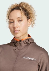 Hnedá zapínacia mikina s kapucňou a oranžovým detailom na golieri, s bielym logom "TERREX". Má textúrovanú tkaninu a štíhly strih.