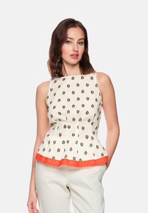 Frau mit schulterlangem braunem Haar, die ein ärmelloses, cremefarbenes Oberteil mit kleinen Blumenmustern und oranger Saum trägt, kombiniert mit hellen Hosen.