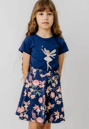 Junges Mädchen mit langen Haaren, das ein marineblaues Hemd und einen Rock trägt, beide mit pinkfarbenen Feen- und Blumenmustern verziert.