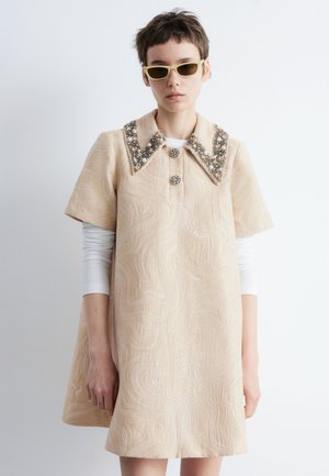 Person mit kurzen Haaren, die ein beigefarbenes, strukturiertes Kleid mit einerperlenbesetzten Kragen, einem weißen Langarmshirt darunter und gelb getönten Sonnenbrillen trägt.