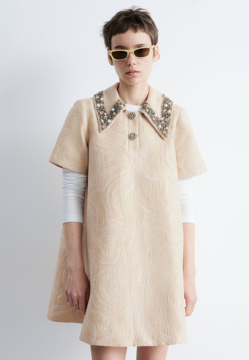 Person mit kurzen Haaren, die ein beigefarbenes, strukturiertes Kleid mit einerperlenbesetzten Kragen, einem weißen Langarmshirt darunter und gelb getönten Sonnenbrillen trägt.