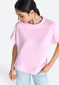 Haut rose clair à manches courtes avec un col rond, coupe décontractée, texture lisse, et un ourlet qui tombe légèrement en dessous de la taille.