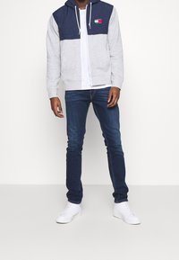 Sudadera con cremallera gris y parte superior azul marino, que presenta un patrón en relieve y un logo. Combinada con jeans azul oscuro y zapatillas blancas.