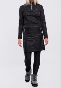 Svart zip-up topp med ett strukturerat mönster, kombinerad med en svart quiltad kjol med sidodragkedjor. Modellen bär svarta leggings och fluffiga stövlar.