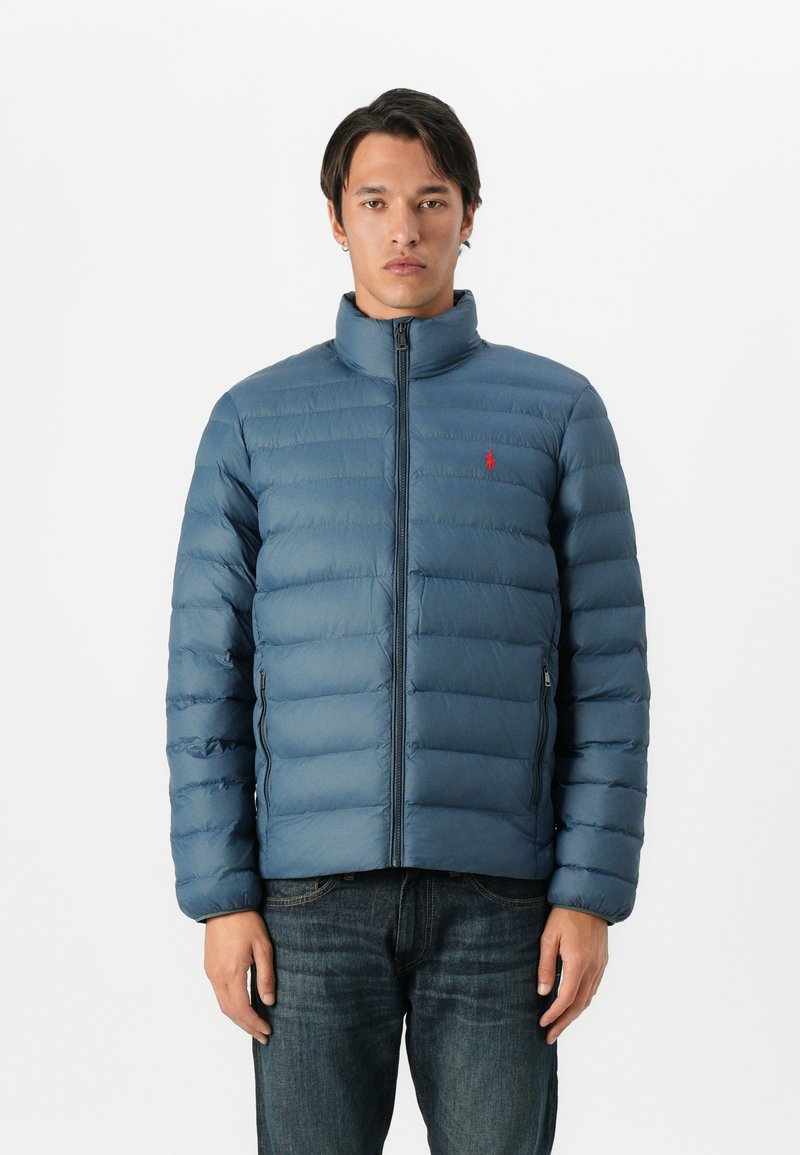 Hombre que lleva una chaqueta acolchada azul con cremallera frontal y un logo rojo, combinada con jeans de mezclilla azul oscuro, de pie contra un fondo blanco plano.