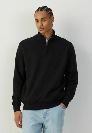 Młody mężczyzna z zaplecionymi włosami, ubrany w czarny półrozpinany sweter i jasnoniebieskie dżinsy, stojący na jednolitym białym tle.