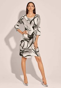 Das schwarz-weiße Kleid zeigt abstrakte Blattmuster, dreiviertel-lange Ärmel, ein Wickeldesign und einen knielangen Saum. Kombiniert mit silbernen High Heels.