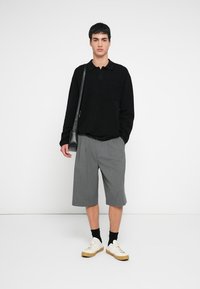 Schwarzes, strukturiertes Langarm-Poloshirt, graue Faltenhose, weiße Sneaker mit beige-farbener Sohle, schwarze Socken und eine schwarze Umhängetasche.