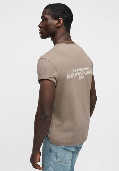 Χακί μπλουζάκι με στρογγυλή λαιμόκοψη και γυρισμένα μανίκια, με κεντημένο κείμενο στην πλάτη. Το ύφασμα είναι απαλό με λεία υφή. Φοριέται με ανοιχτόχρωμο μπλε τζιν.