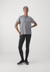 Šedé tričko s krátkými rukávy, volný střih; kombinováno s černými legínami; model má na sobě černé tenisky. Hladký povrch, minimální detaily.