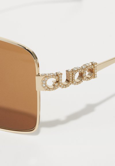 Zbliżenie na złote okulary przeciwsłoneczne z brązowymi soczewkami, ozdobione koralikowym logo "Gucci" na ramieniu, na białym tle.