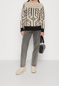 Stickad tröja i krämfärg med svarta geometriska mönster, kombinerad med grå jeans och beige plattformstövlar. Håller en svart handväska.