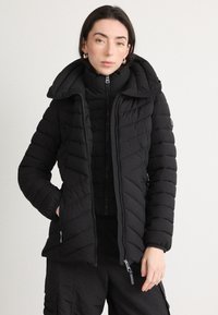 Giacca a vento nera con colletto alto e cappuccio, motivo a rombi, cerniere doppie e tasche laterali. Realizzata in materiale leggero e isolante.