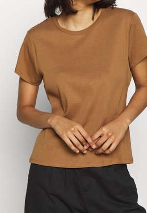 Nainen, jolla on päällään ruskea lyhythihainen kauluspaita ja mustat housut, kädet löyhästi vyötärötasolla ristissä.