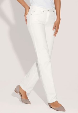 Vrouw die witte rechte jeans en lichtgrijze lage schoenen met puntige neus draagt, staand met gekruiste benen.