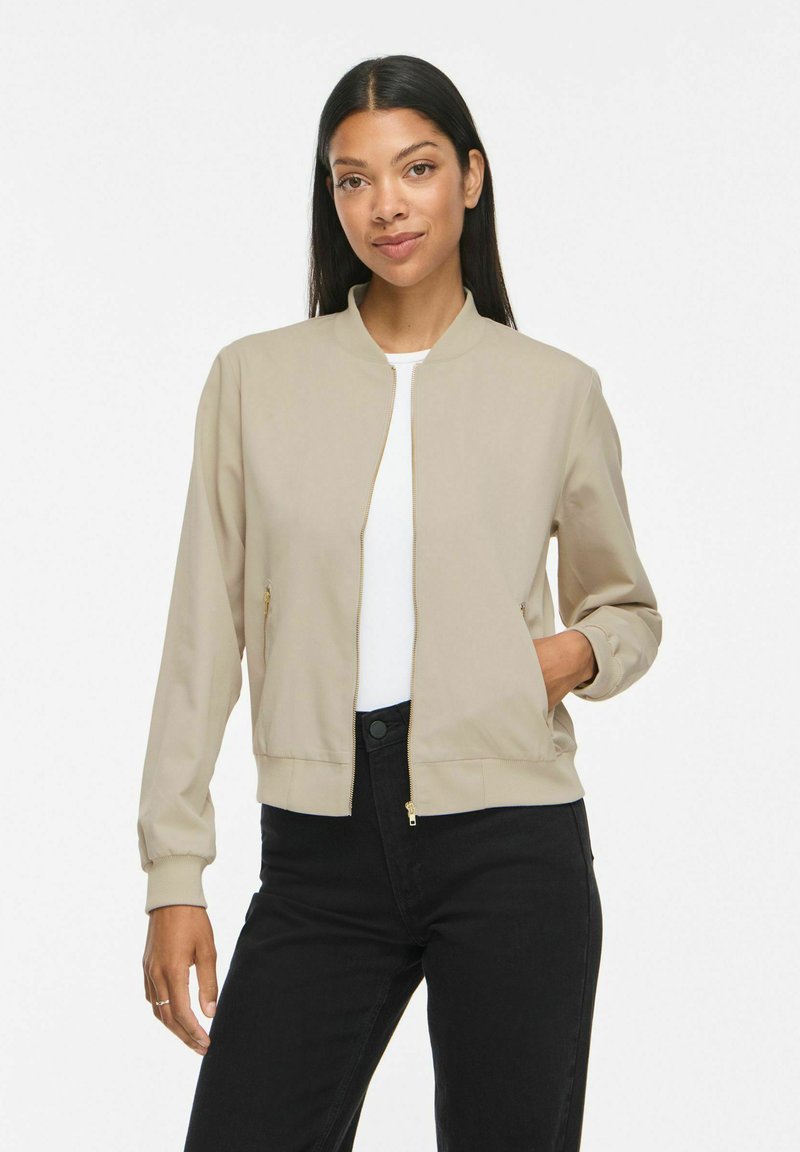 Femme aux longs cheveux foncés portant une veste beige, une chemise blanche et un pantalon noir, debout avec une main dans la poche de sa veste.
