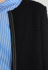 Černá texturovaná bunda se stříbrným zipem, kombinovaná s světle modrou a bílou proužkovanou košili. Důraz na látku a kontrastní barvy.