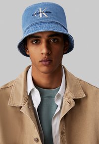 Blå denim bucket hat med broderet logo, iført med en beigemantel over en grøn sweater og en hvid skjorte med krave. Portræt i nærbillede.