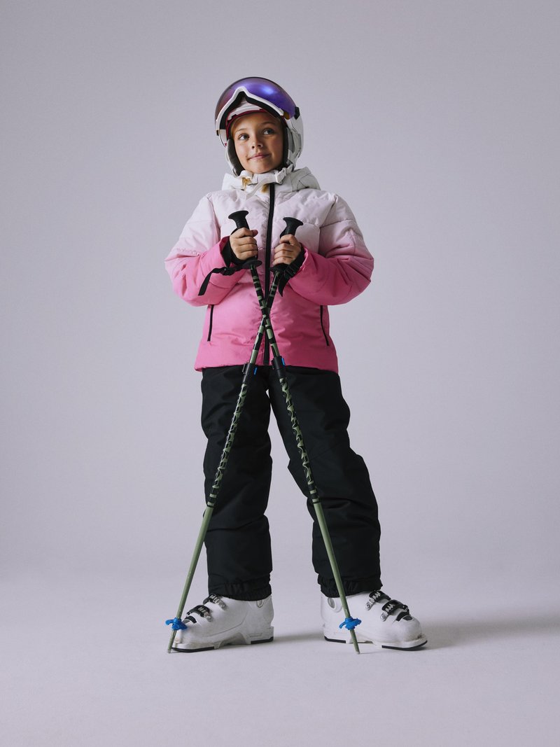 Skioutfit bestående av en rosa och vit jacka, svarta byxor, vita skidstövlar och skidstavar. Tillbehör inkluderar en vit hjälm och spegelglasögon.