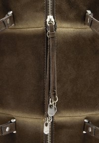 Brązowa zamszowa tkanina z centralnym srebrnym zamkiem błyskawicznym z dwoma uchwytami oraz ozdobnymi zamszowymi frędzlami, otoczona skórzanymi paskami z srebrnymi ćwiekami.