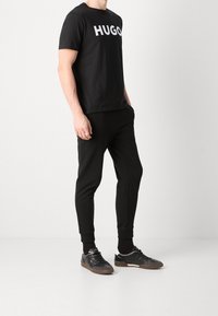 Camiseta negra con el logo "HUGO" en blanco, combinada con pantalones jogger negros y zapatillas deportivas negras con suela marrón. Diseño simple y ajustado.