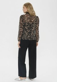Durchsichtige schwarze Bluse mit braunem Tigerdruck, lange Ärmel und hohem Kragen; kombiniert mit lockeren schwarzen Hosen und silbernen Absatzschuhen.