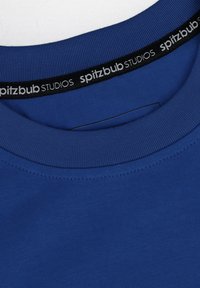 Blaues T-Shirt mit Rundhalsausschnitt, geripptem Kragen und einem Etikett mit "spitzbub STUDIOS" in Weiß auf schwarzem Hintergrund. Glatter Stoff.