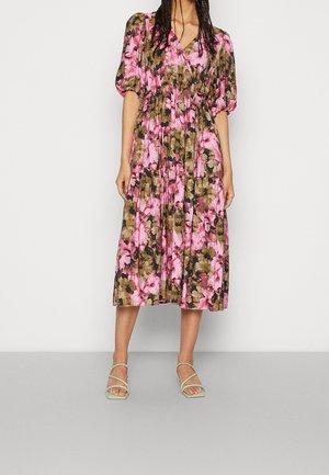 Woman wearing a midi floral dress with pink, brown, and black colors, puff sleeves, and light green strappy sandals standing against a plain background.