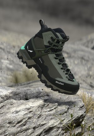 Grøn og sort Salewa vandrestøvle med kraftig sål, snørebånd og Gore-Tex mærke, der hænger over klippefyldt terræn med små planter.