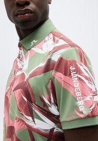 Κοντινό πλάνο ενός ατόμου που φοράει ένα πόλο πουκάμισο με πράσινο και ροζ φλοράλ σχέδιο, με κεντημένο το "J.LINDEBERG" στο μανίκι και στο γιακά.