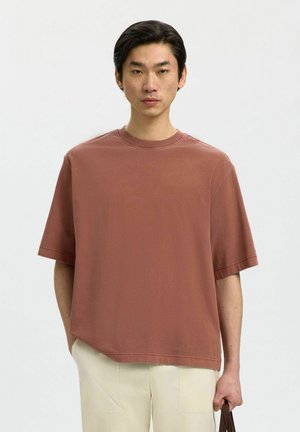 Giovane uomo con capelli scuri che indossa una maglietta larga color ruggine e pantaloni beige chiaro, che tiene con una mano la maniglia marrone di una borsa.