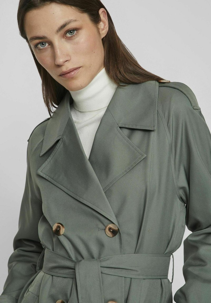 Lichtgroene trenchcoat van gladde stof, met grote knopen, een gestrikte taille en brede revers. Gelaagd over een witte coltrui.