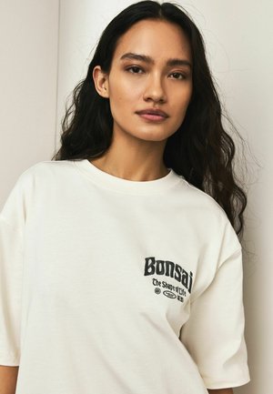 White cotton t-shirt with a relaxed fit, featuring a black printed logo "Bonsai" and "The Shape of Life" on the chest. Soft texture.