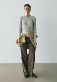 Frau steht vor einer schlichten Wand und trägt ein graues Langarmshirt, braune Hosen mit weitem Bein, eine beige Jacke, die um die Taille gebunden ist, und eine Anhänger-Halskette.