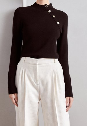 Femme portant un haut noir ajusté à manches longues avec des boutons argentés sur l'épaule et un pantalon large texturé blanc taille haute.