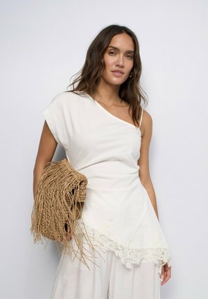 Femme portant un haut blanc asymétrique et une jupe, tenant un sac tissé marron à franges sur un fond clair uni.