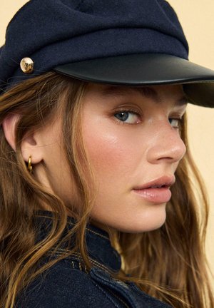 Jonge vrouw met een lichte huid en blauwe ogen, droeg een marineblauwe pet en gouden oorring, met golvend bruin haar en spijkerjas.