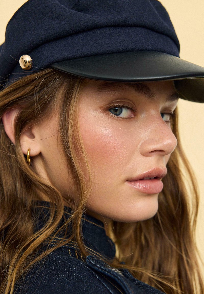 Jeune femme à la peau claire et aux yeux bleus, portant une casquette bleu marine et une boucle d’oreille en or en forme de créole, avec des cheveux bruns ondulés et une veste en jean.
