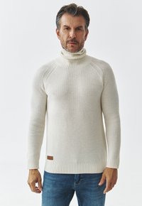 Pulover tricotat cu guler înalt, de culoare alb murdar, cu textură striată și design ajustat. Prezintă un accent din piele la tiv și mâneci lungi.